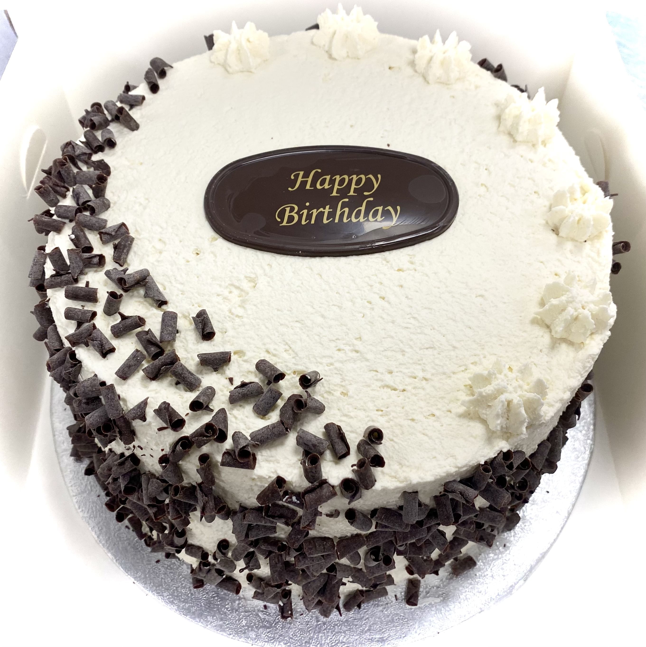 A round, frosted Black Forest birthday cake with white cream topping and edges adorned with chocolate shavings. At the center, there's an oval chocolate plaque with 'Happy Birthday' written in yellow. The cake, infused with cherry liqueur, sits on a silver base, radiating pure chocolate goodness.